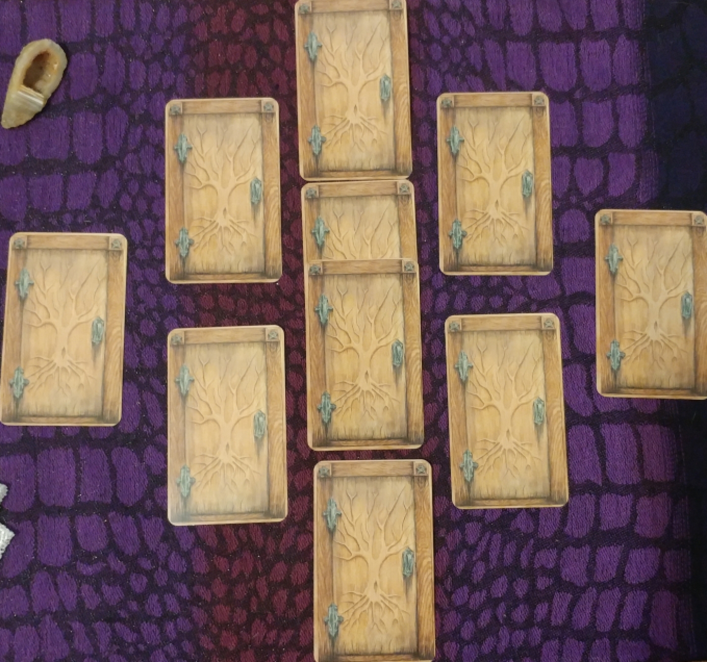 Ten matching cards (faced-down) that resemble unopened wooden doors, hinting at the unknown. The frame of each door is of a darker wood, two pentagrams etched in the top corners. A naked tree is etched into the door with the roots reaching down towards the earth, the branches reaching towards the sky; this encompasses the three realms of the deep influences of the past, the present moment, and the influences from above. The cards are arranged to resemble the vague shape of a cauldron (including a handle) with two extra cards inside the cauldron layout. There is a tawny-colored banded agate druzy to the left of the spread.