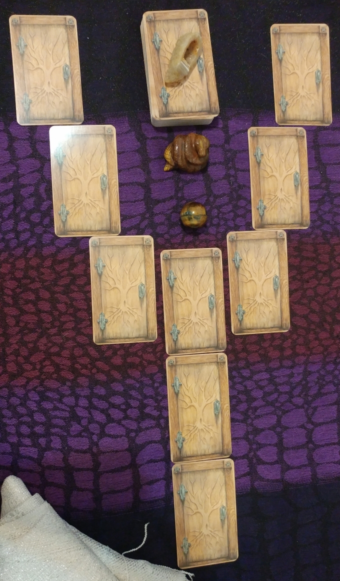 Nine matching cards (faced-down) that resemble unopened wooden doors, hinting at the unknown. The frame of each door is of a darker wood, two pentagrams etched in the top corners. A naked tree is etched into the door with the roots reaching down towards the earth, the branches reaching towards the sky; this encompasses the three realms of the deep influences of the past, the present moment, and the influences from above. The cards are arranged as a two-tined fork. There is a tawny- colored banded agate druzy on the remainder of the deck, which is sitting between the two forking paths. There is also a coiled snake carved from tiger's eye and a sphere of hawk's eye in the fork. A silvery-white scarf accents the bottom left corner of the picture.