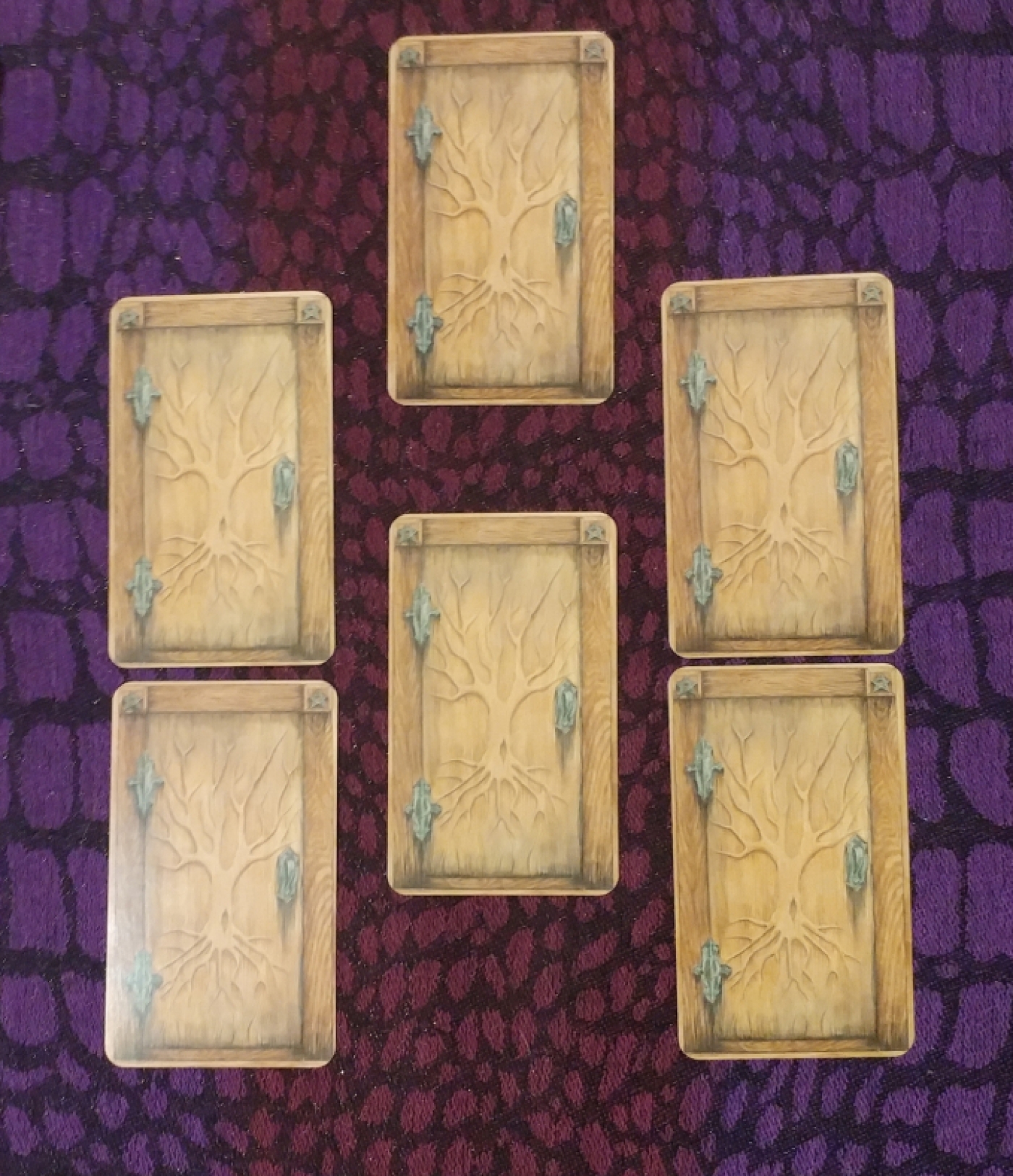 Six matching cards (faced-down) that resemble unopened wooden doors, hinting at the unknown. The frame of each door is of a darker wood, two pentagrams etched in the top corners. A naked tree is etched into the door with the roots reaching down towards the earth, the branches reaching towards the sky; this encompasses the three realms of the deep influences of the past, the present moment, and the influences from above. The cards are arranged to resemble the points of a pentagram (five-pointed star comprised of the elements earth, air, water, fire, and spirit) with an extra card in the center.