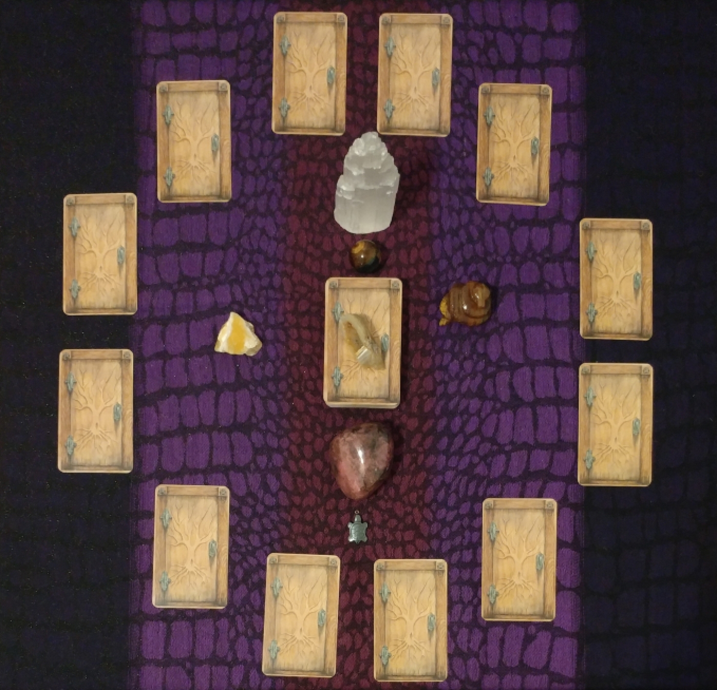 Twelve matching cards (faced-down) that resemble unopened wooden doors, hinting at the unknown. The frame of each door is of a darker wood, two pentagrams etched in the top corners. A naked tree is etched into the door with the roots reaching down towards the earth, the branches reaching towards the sky; this encompasses the three realms of the deep influences of the past, the present moment, and the influences from above. The cards are arranged in a circle. The remainder of the deck lays in the center. There is a tawny-colored banded agate druzy on top of the deck. Other gemstones are surrounded by the wheel (raw selenite tower, raw yellow calcite, polished hawk's eye orb, coiled tiger's eye snake, polished rhodinite, and hematite turtle amulet).